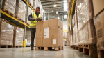 Mid-market warehouse with pallets of furniture cartons; worker scans shipment to improve purchase order management accuracy.